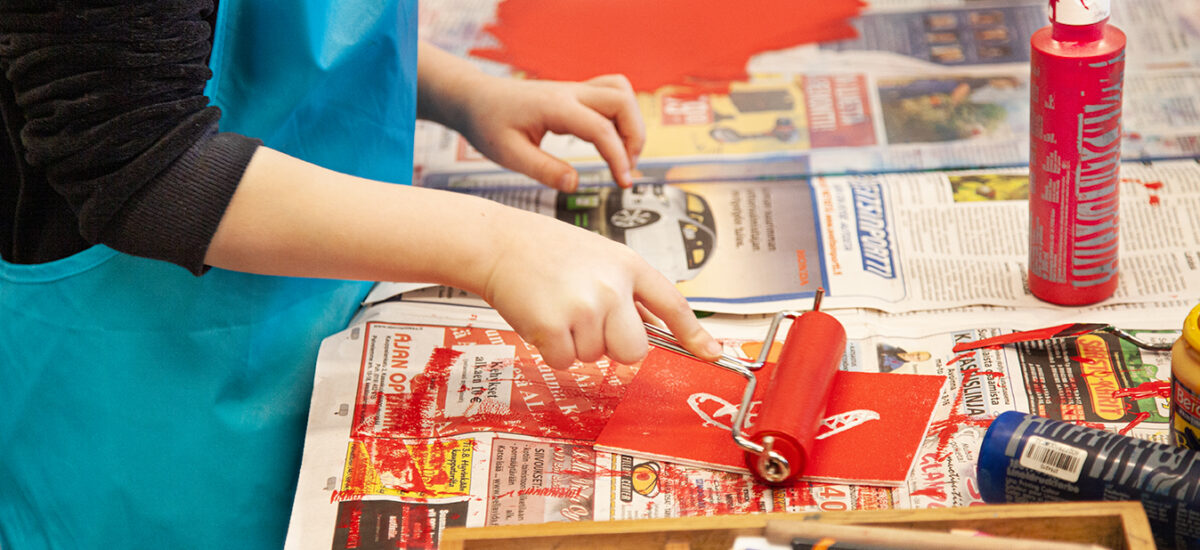 Lähikuva sanomalehdillä vuoratusta pöydästä, jonka ääressä turkoosiin essuun puheutunut lapsi rullaa punaista väriä telalla grafiikkaprinttiin